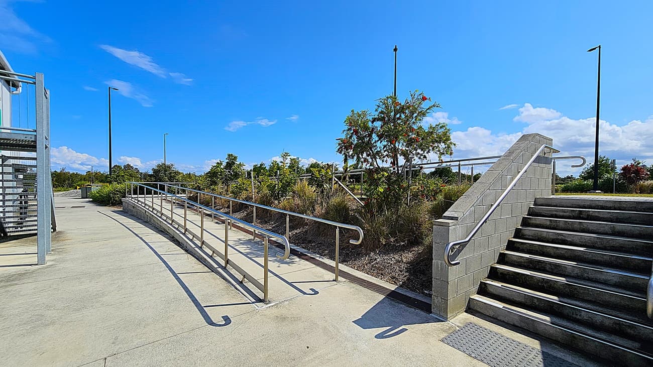 On the left, a set of concrete steps leads up to a building with dark doors, flanked by a small tree. The steps are equipped with metal handrails for support. To the right, a wide, smooth concrete pathway curves away, bordered by a strip of grass. The building features a flat roof supported by columns, with large windows visible along its length.