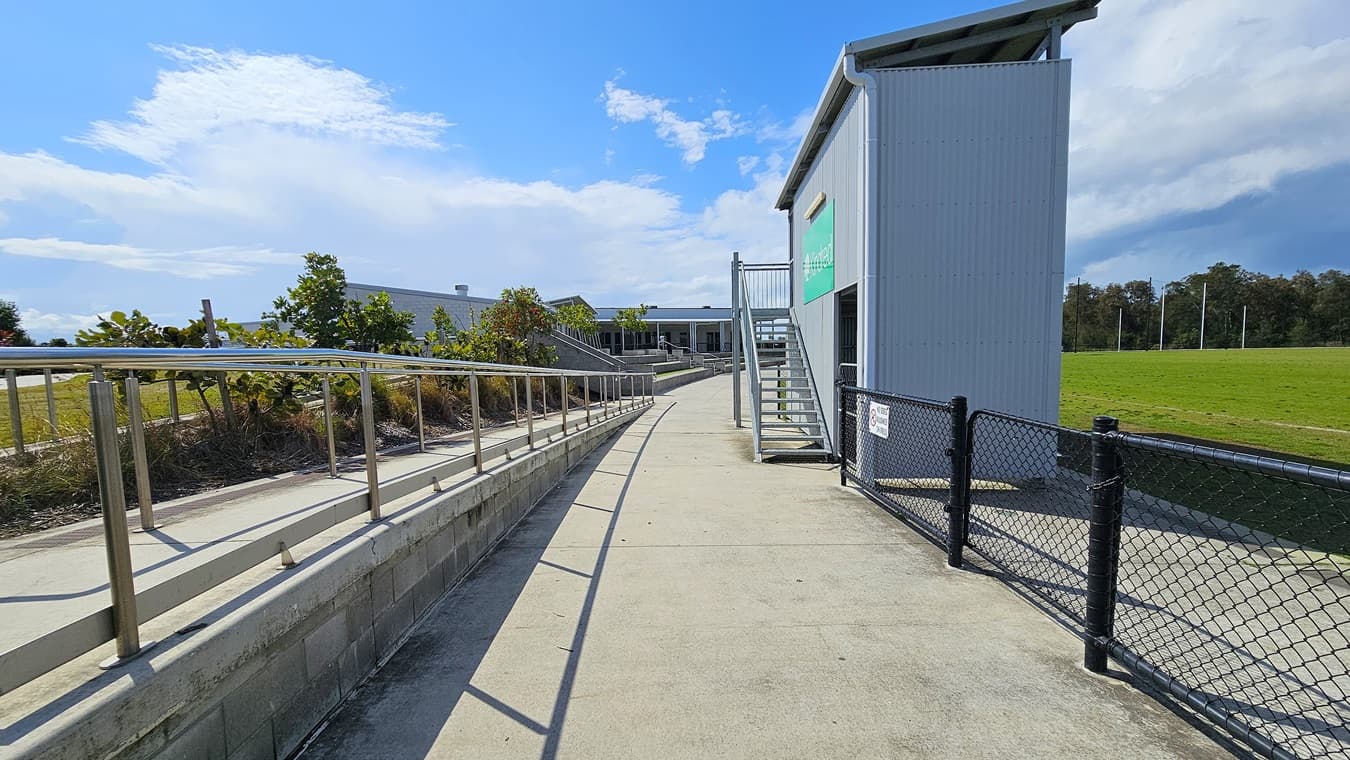 On the left, a gently sloping concrete ramp with a metal handrail leads towards a grassy area, bordered by low shrubs. The surface is smooth and grey, extending towards a building in the background. To the right, a set of metal stairs ascends to a higher level of the building, which has a grey exterior and a green sign. A black chain-link fence runs along the pathway, separating it from the adjacent field.