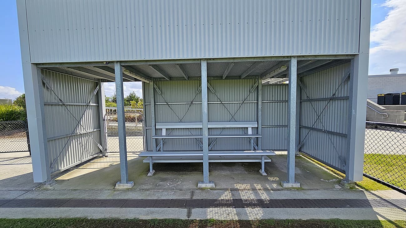 On the left, a grey metal structure features a covered seating area with two long benches, supported by vertical metal posts. The benches are positioned against a backdrop of corrugated metal walls. To the right, a chain-link fence encloses the area, providing a boundary. The floor is a combination of concrete and grass, with a smooth concrete pathway leading up to the seating area. The ceiling is flat and made of metal, matching the walls, creating a sheltered space for spectators.