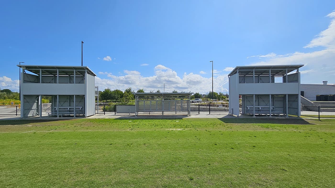 On the left, a grey metal structure features a covered viewing area with benches, supported by vertical posts. Adjacent to it, a similar structure is visible on the right, both facing a grassy field. In the middle, a concrete pathway runs between the two structures, leading towards a fenced area in the background. The floor is primarily grass, with some bare patches, and the ceiling is flat and made of metal, providing shelter for spectators.