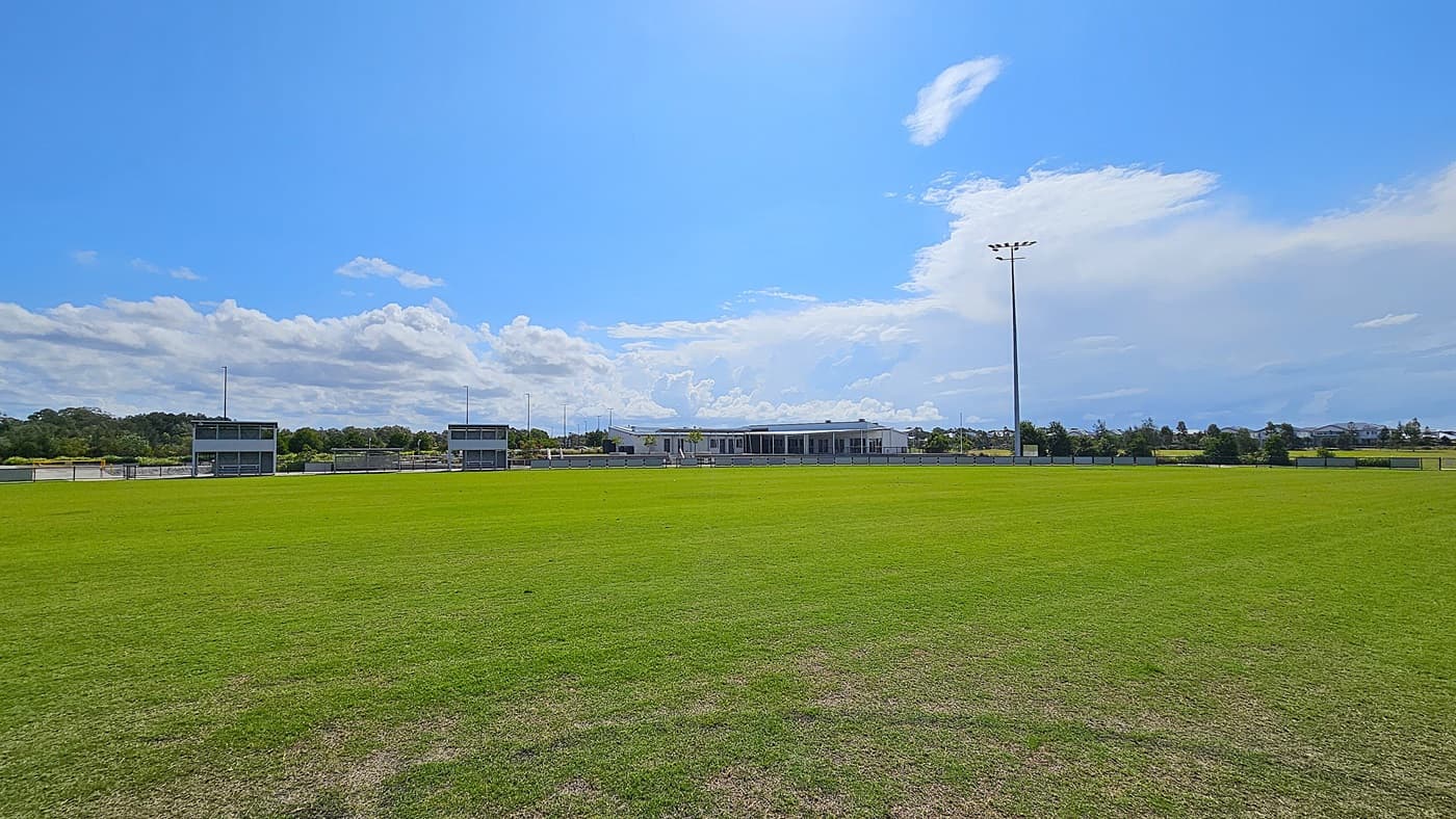 On the left, two small grey structures with large windows serve as viewing areas, each with a set of steps leading up to them. To the right, a larger building with a flat roof and multiple windows is visible, positioned further back from the field. The foreground features a well-maintained, expansive green grass field, while the background includes trees and additional structures.