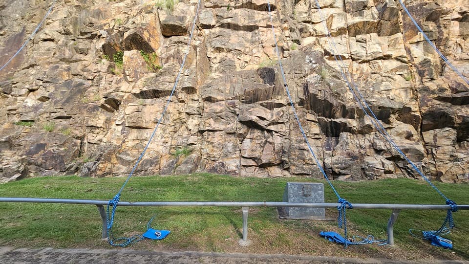 Five blue ropes hang from the cliff, fastened to a metal railing near the road. The cliff has various shaped brown rocks with both smooth and sharp edges, and wild grass grows beneath them. At the base of the cliff is a grassy area, with a solid concrete structure bearing a light fixture positioned on the grass.