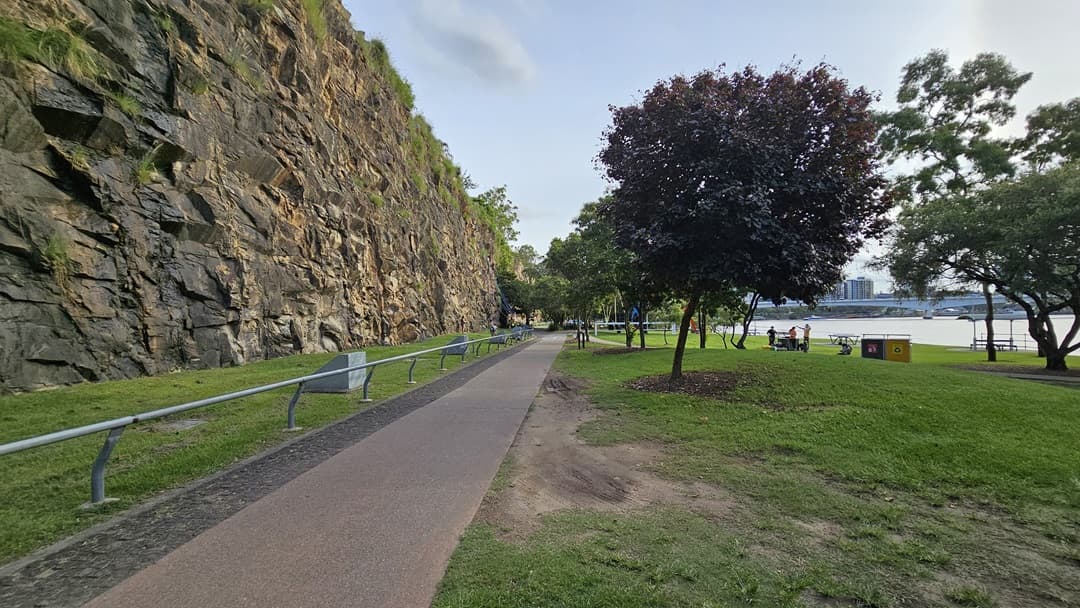 On the left is a cliff overlooking a straight road with a metal railing. Concrete structures with a light fixture stands on the grass nearby. To the right of the road is the river and an open space with green grass, trees, picnic tables, and garbage bins.