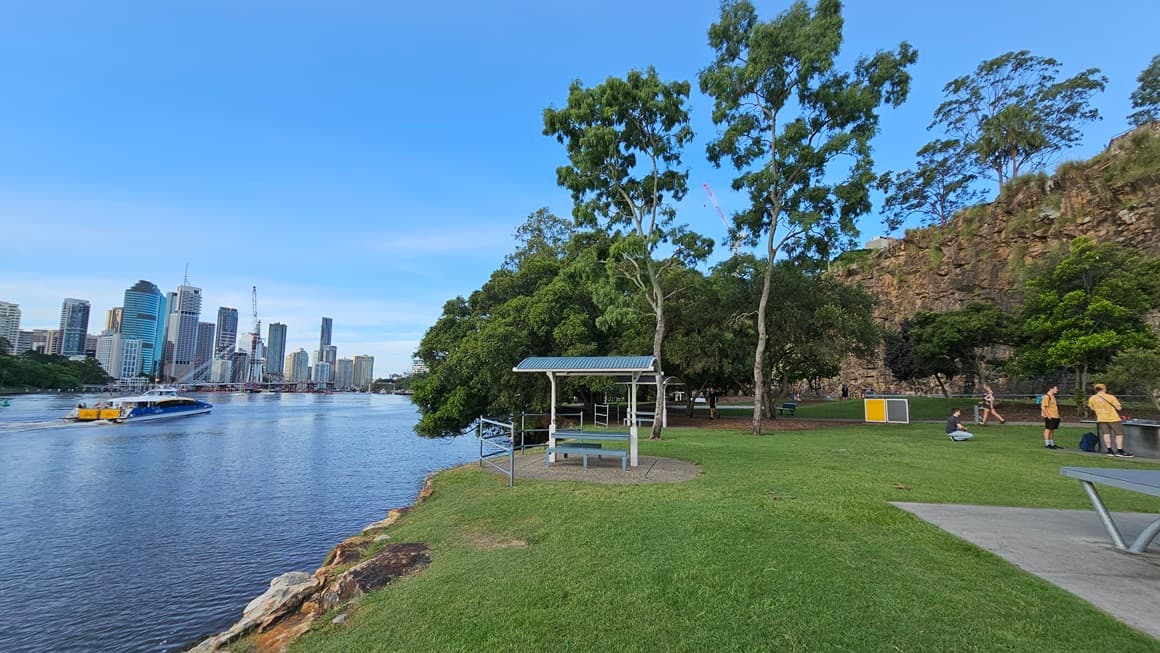 On the left is the river with a ferry boat, while skyscrapers form the backdrop. To the right is a spacious green area with picnic tables, benches under tree shades, and tall trees surrounding the space. Trash bins are visible. On the far right is a cliff. People occupy the space and move about.