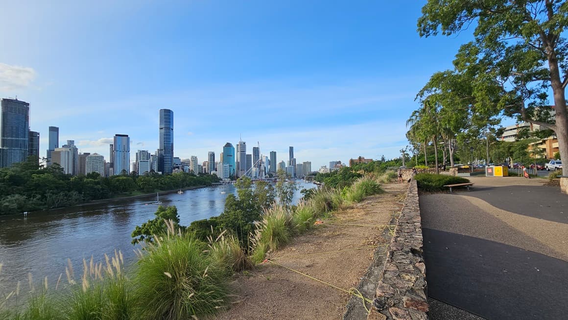 On the right is a park with bench chairs positioned under trees and landscaped plants. A concrete bench barricades the area. The road is on the right with vehicles, and a multi-story building is visible in the distance. To the left, the river flows below, with wild grass and trees covering the cliff. Skyscrapers and trees fill the backdrop.