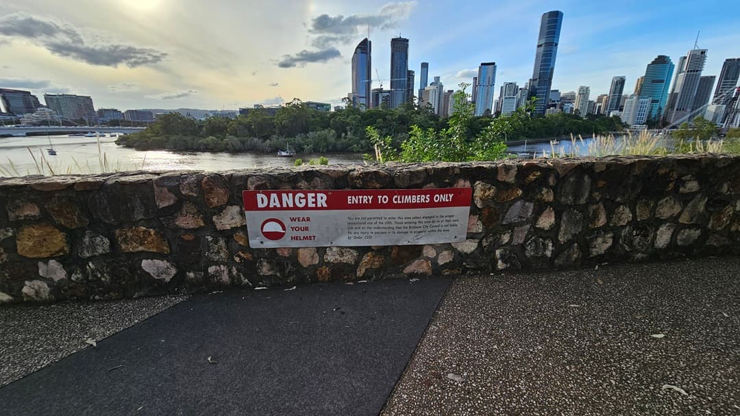 A panoramic view of the river from the cliff top. Several ferry boats navigate the river. Trees and green grass line the riverbank, while skyscrapers dominate the backdrop. The cliff is covered with trees and wild grass. A concrete barrier with a warning sign borders the road. The flooring consists of pebblestone and black concrete.