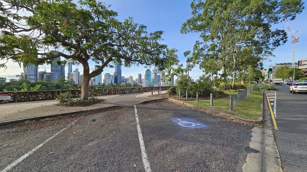 An accessible parking space with markings. Next to it is a landscaped area with green grass, tall trees, and ornamental plants, bordered by concrete bollards. A bench chair is positioned nearby. To the right is a road with traffic lights, cars, buildings, and a tower crane. Skyscrapers are visible on the far left.