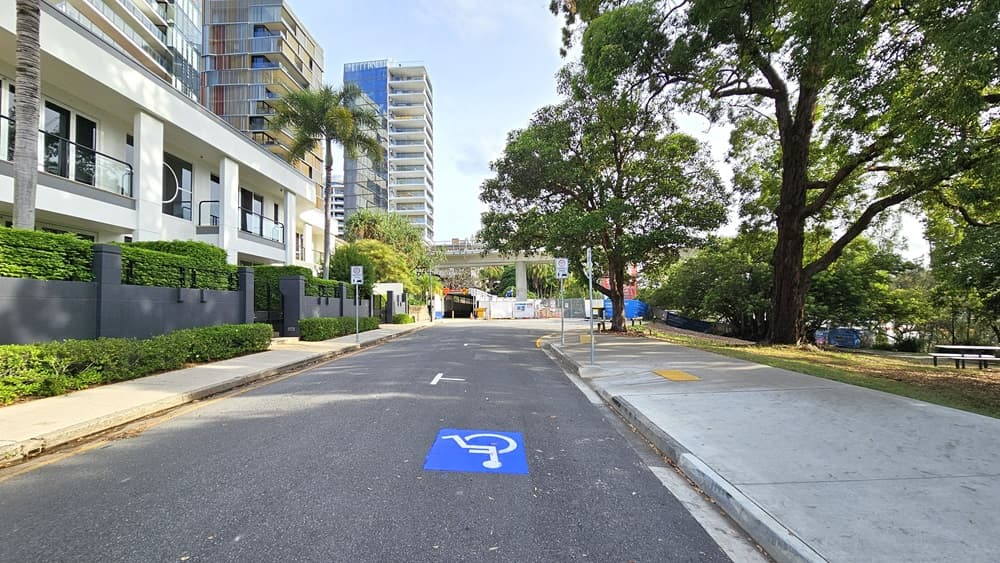 To the left is a multi-storey building with a grey wall and landscaped plants in front. An accessible parking sign is on the road. Road signs are also positioned in front of the building gate. A step-free ramp leads to a slightly elevated pathway. Trees and grass border the right side. At the end, the road curves right.