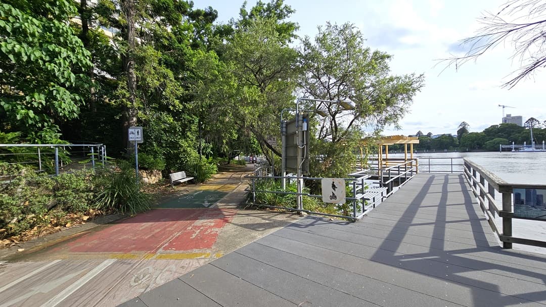 The road divides, with the right path leading to a viewing deck overlooking the river. The left path, marked with red and green, continues straight ahead. Bench chairs are positioned under the shade of trees, and a metal fence borders the road.
