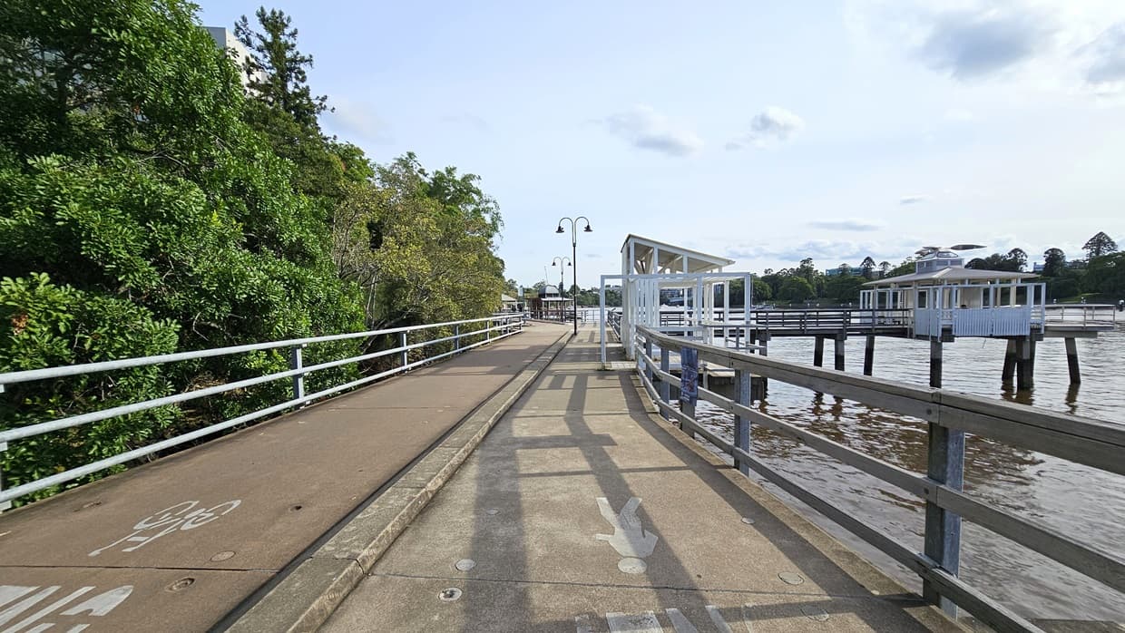 A concrete median divides the road: the left side for bicycles and the right for pedestrians. Metal railings and trees border the left side, while the Brisbane River is to the right. A ramp leads to a shaded structure on the right. Tall street lights line the road.
