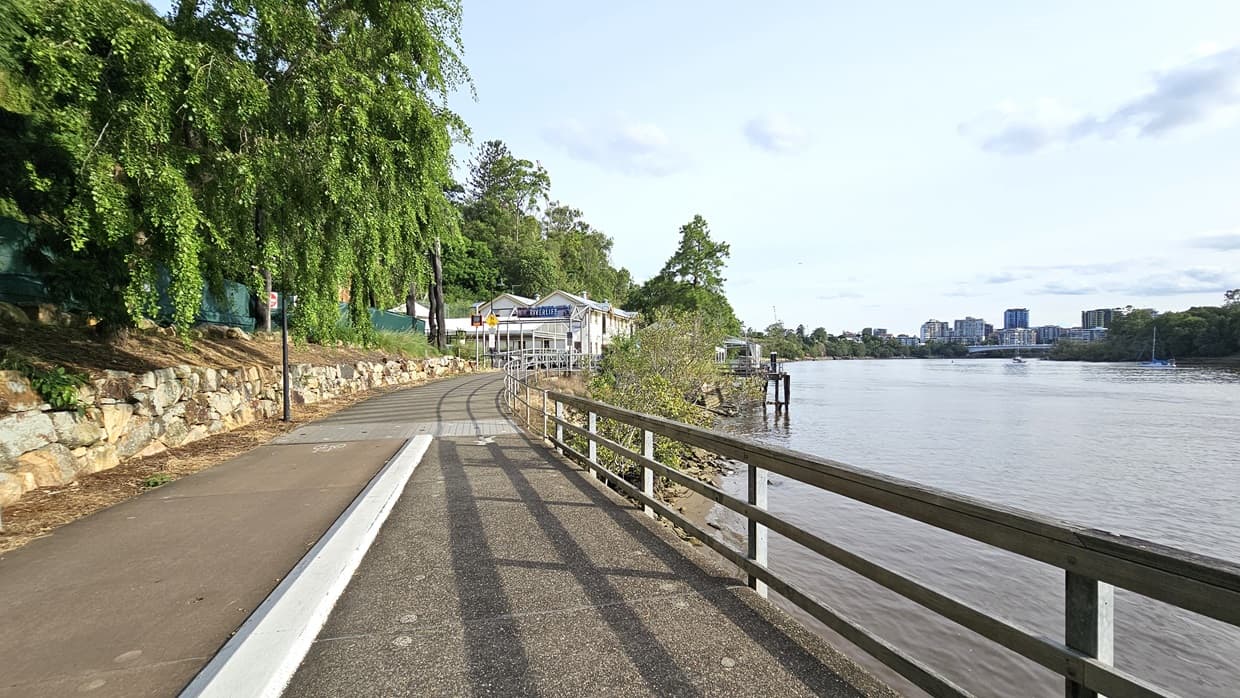 A concrete median divides the road: the left side for bicycles and the right for pedestrians. Large rock formations and green mesh, trees border the left side, while the Brisbane River is to the right. Metal railings line the road on the right. Further ahead, the Riverlife facility building is visible.