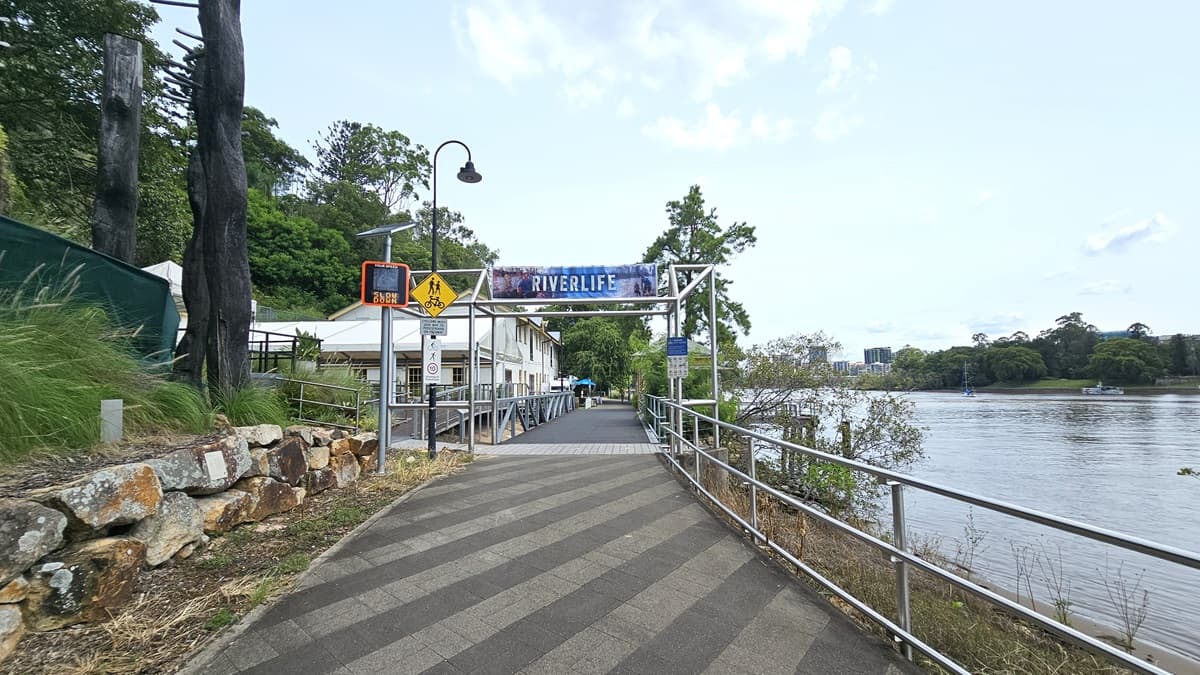 In the centre stands a metal arc displaying the facility name. A lamppost with signages is on the left along with a solar-powered digital signage. Rock formations, wild grass, green mesh, and trees border the left side, while the Brisbane River is to the right. Metal railings line the road on the right. A ramp with handrails leads to the building. The road has a mix of white and grey bricks.