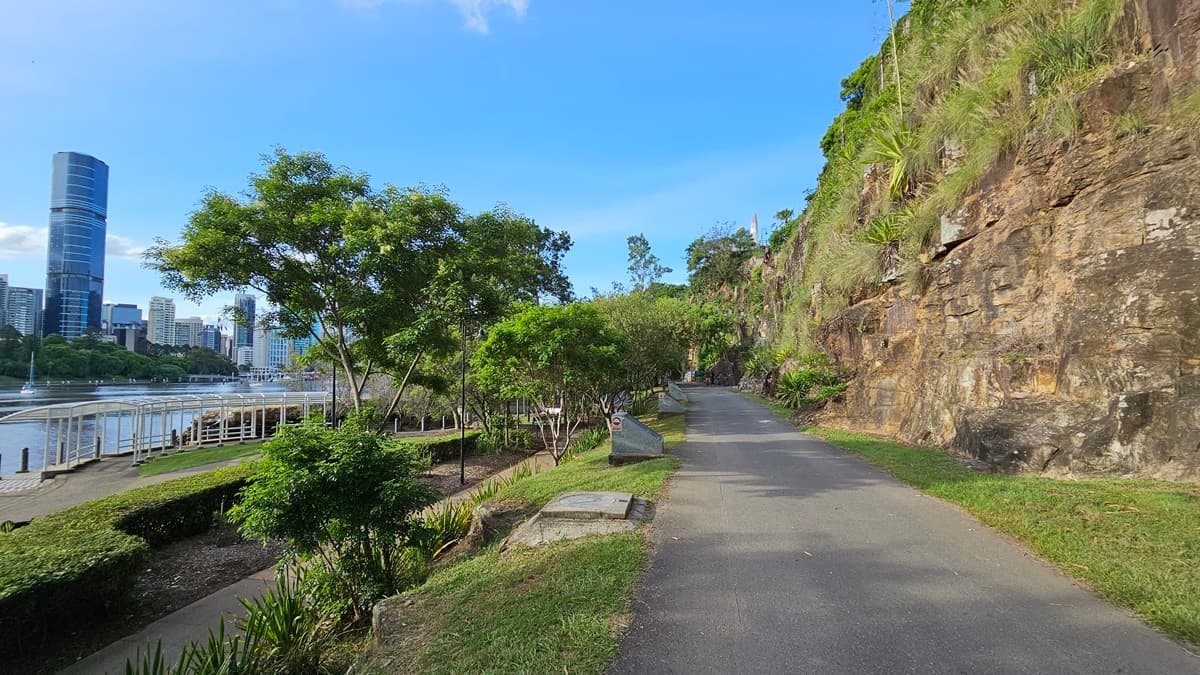 A straight road is bordered by a cliff on the right and trees and solid structures with lighting on the left. To the left of the road is lower ground with landscaped plants and neatly trimmed shrubs. Shade structures are positioned near the lake. Further ahead, tall skyscrapers are visible.