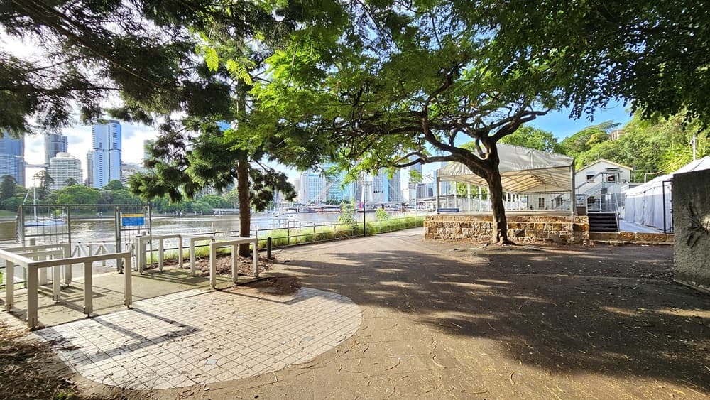 To the right is an open-shaded structure with steps. A wall with a fishbone design is nearby. On the left is an entrance to the ramp equipped with metal railings. The entrance to the ramp has grey tiles. Tall buildings are visible on the other side of the river, while tall trees border the road.