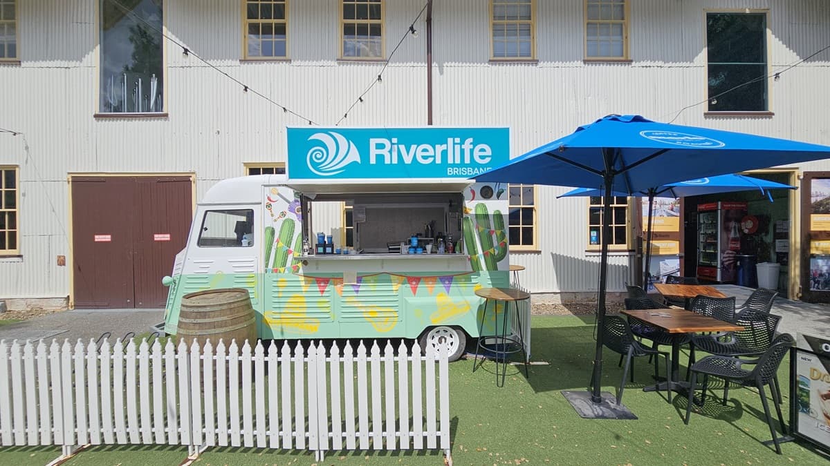 The cafe counter resembles a mini bus, painted green with tropical designs and facility signage on top. A barrel and a low white fence decorate the front. Movable tables and chairs are arranged under large blue umbrella shades. To the right is the entrance to the reception building, with a fridge displaying cold drinks visible. Behind is a white, two-storey building with a corrugated facade. Synthetic turf covers the ground.