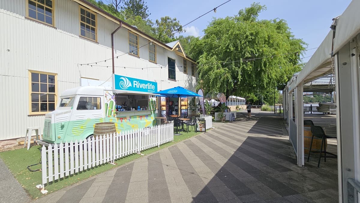 The cafe counter resembles a green mini-bus with tropical designs, featuring the facility name on top. A barrel and low white fence adorn the front. Movable tables and chairs are arranged under large blue umbrella shades. Behind is a two-storey white building with a corrugated facade. Synthetic turf covers the ground on the left, while the road on the right has grey bricks. A venue tent with chairs underneath is on the right.
