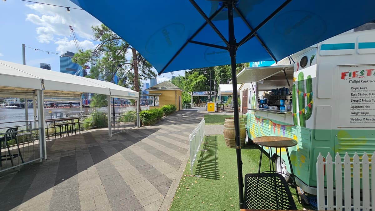 On the right is a cafe counter resembling a green mini-bus with a tropical design. A barrel and low white fence adorn the front. Movable tables and chairs are arranged under large blue umbrella shades. Synthetic turf covers the ground on the right, while the road on the left has grey bricks.  On the left is a venue tent with tables and chairs underneath. The river and tall skyscrapers are visible in the background.