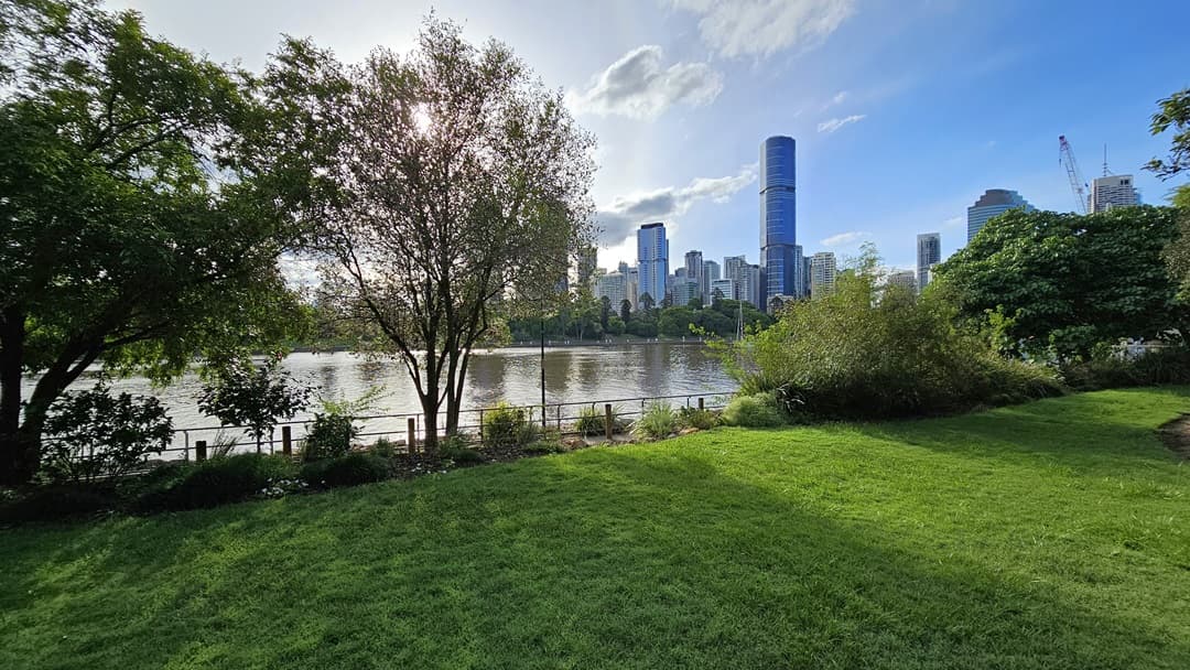 An open green space with neatly trimmed grass beside the river with trees and plants lining both sides. A tall lamp post stands near the road, adorned with red triangle banners. Trees are also seen along the riverbank, while skyscrapers form the backdrop.