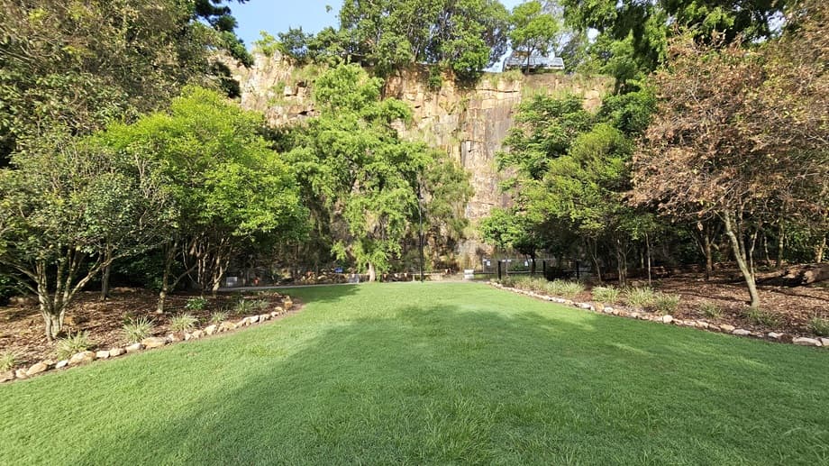 An open green area with well-maintained grass bordered by rocks and trees on both sides. In the centre is a cliff surrounded by trees and wild grass.