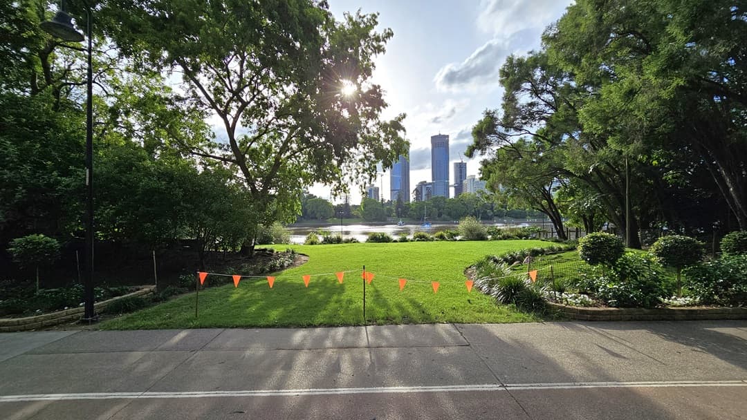 A well-maintained green area beside the river, with neatly trimmed grass and trees on both sides. A tall lamp post is situated near the road, decorated with red triangle banners. Trees line the riverbank, with skyscrapers in the background.