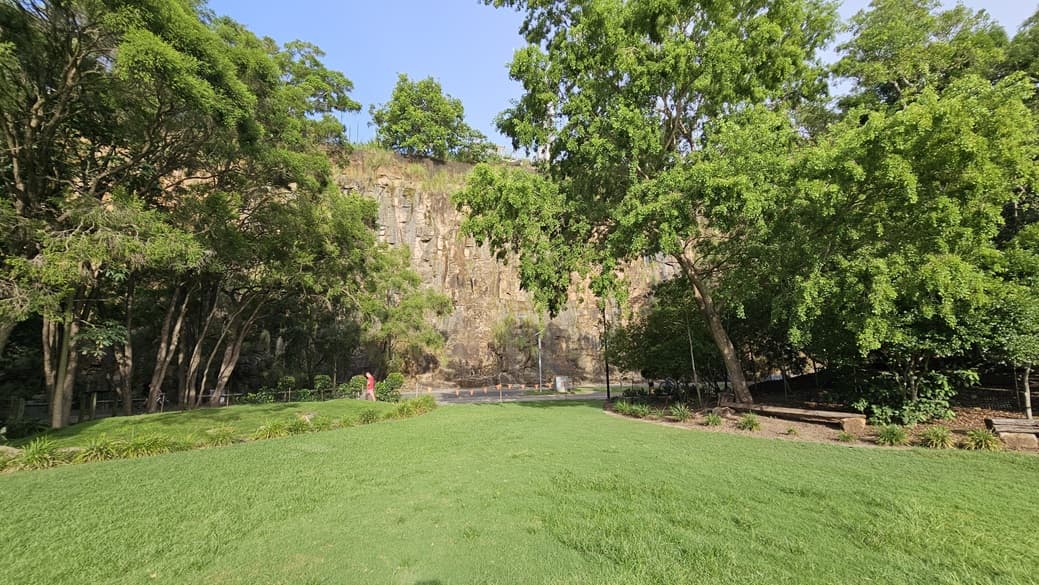 A serene green space with neatly kept grass, flanked by ornamental plants. Bench chairs are placed under the shade of tall trees on either side. In the centre, a cliff is surrounded by trees and wild grass. Red triangle banners are hung across to mark the boundary of the area.