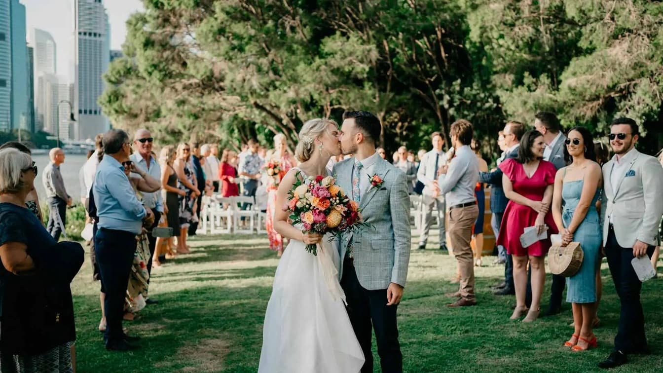 An image showing how the venue looked during a wedding event. In this image, the bride, holding a wedding bouquet, and the groom stands in the middle, guests surrounding them on the grassy area. Tall skyscrapers and tall trees fill the background.