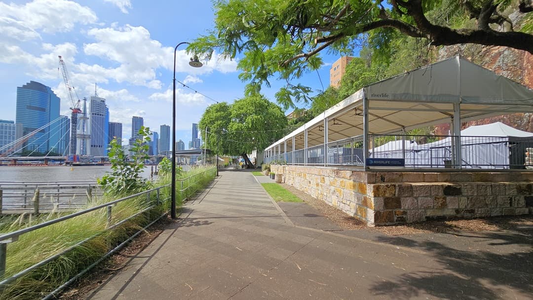 On the left stands a venue space with a pitched white tent roof and metal railings. The outer wall has brown bricks. To its left is an area with synthetic turf, a table, and potted plants. Behind the tent are grey buildings, while trees and wild grass cover the cliff in the background. To the right is a straight road with tall lamp posts and the river. Trees, wild grass, and a metal fence border the road on the right.