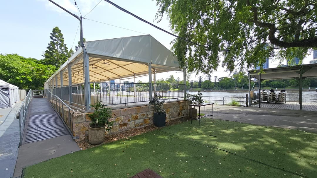 A venue space with a pitched white tent roof and metal railings. The outer wall has brown bricks. To its left is a step-free ramp with timber floor and metal railings. Adjacent is an area with synthetic turf, a table, and potted plants. A shaded structure with timber floor stands in front of the river, with kayaks arranged underneath. Trees, wild grass, and a metal fence border the road on the right.