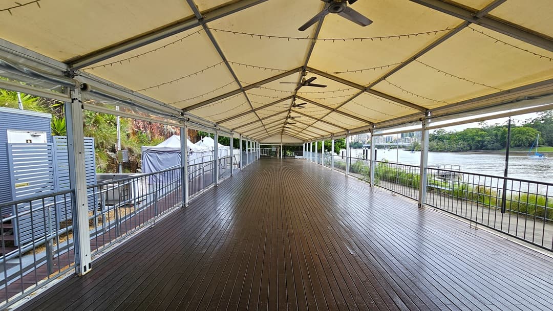 The venue space features a long open area with a pitched tent roof, illuminated by light fixtures and a ceiling fan. The floor has polished timber and metal railings surrounding the space. On the left is a cliff with wild grass and plants, alongside other tents and grey buildings. On the right is the river and the road, bordered by trees and wild grass.