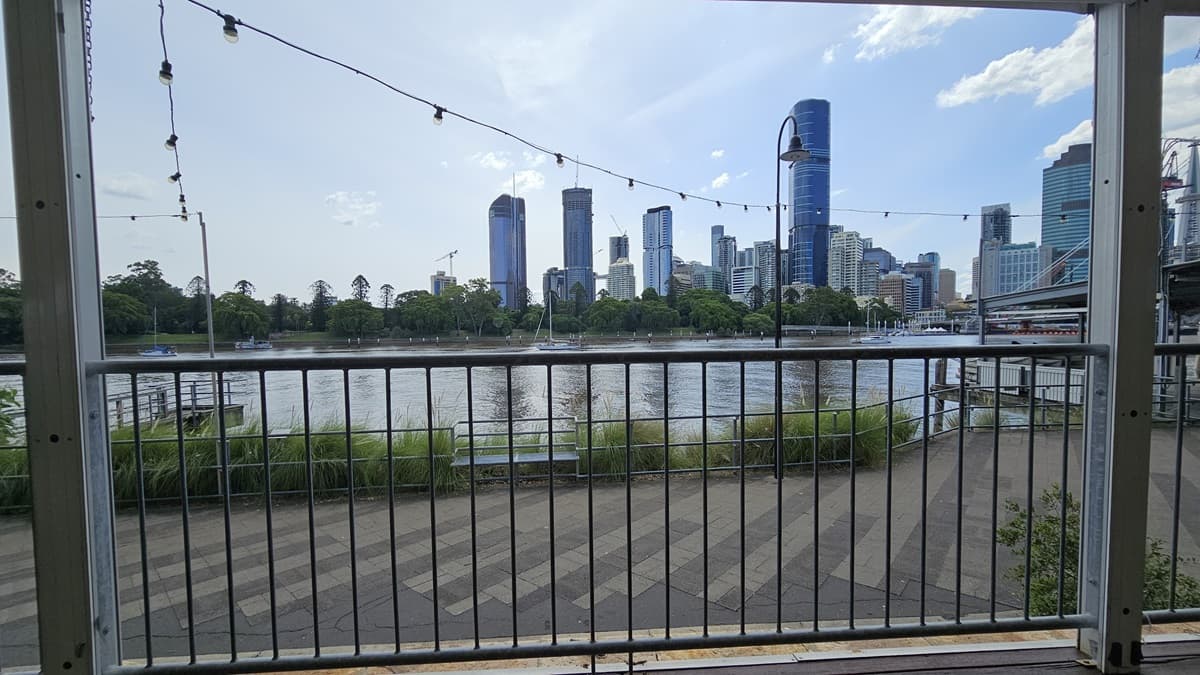 Metal railings secure the space with overhead light fixtures. Ahead lies the road and the river, bordered by metal railings and wild grass. A bench chair sits at the road's edge. A section of the ramp is visible to the right, while a shaded structure store kayaks to the left. Skyscrapers, trees, and the bridge adorn the backdrop.