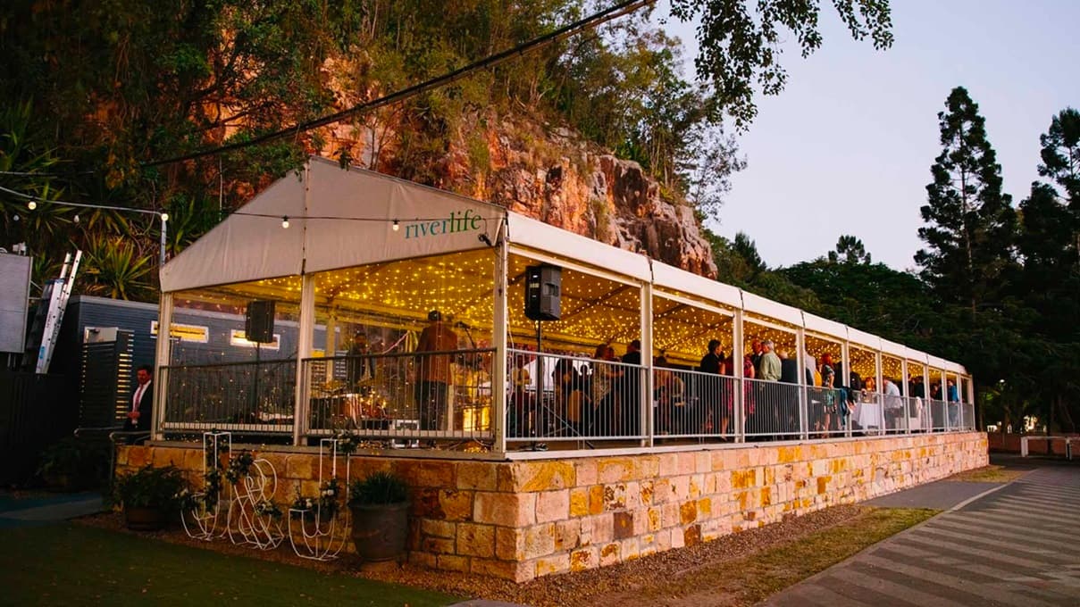 The image depicts a venue bustling with guests during an event. Metal fencing encloses the space while light fixtures adorn the ceiling. Audio speakers are strategically placed in the corners. On the left, the cliff, additional tents, and grey structures can be seen. The tent's outer wall is made of grey-brown bricks adorned with potted plants and lettering. Trees and wild grass complete the scene.
