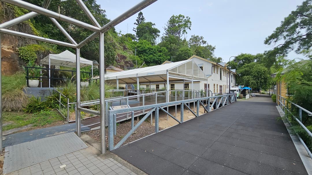 On the left, a step-free ramp with metal handrails and truncated domes leads to a venue tent. Nearby, more venue tents stand close to the cliff. To the right, a straight road is equipped with railings. At the end of the railing is a building with a cream facade. Further along, a two-storey white building is positioned on the left side of the road. Trees and wild grass border the road and the cliff on the left.
