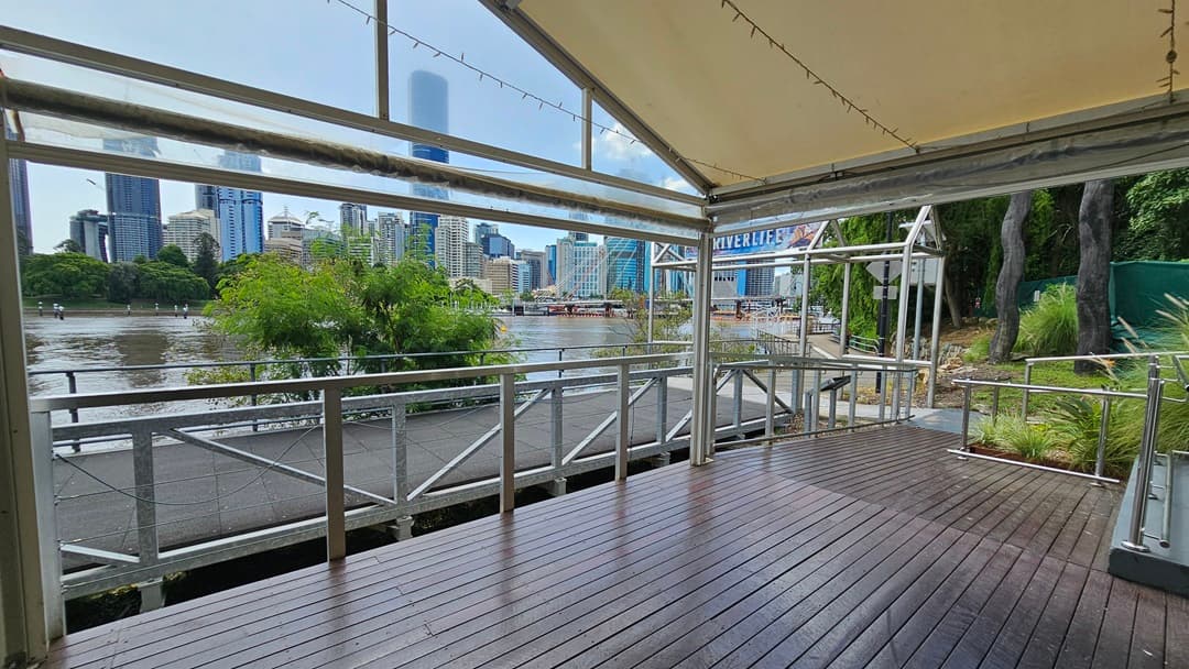 On the right, a step-free ramp with handrails leads to the venue tent. The tent features metal railings and timber floors. To the right, there's greenery with wild grass, tall trees, and green mesh. A metal arc displaying the facility name is also visible. To the left, there's a road and a river. Trees and metal railings border the left side of the road. Skyscrapers and the iconic bridge are visible in the background.
