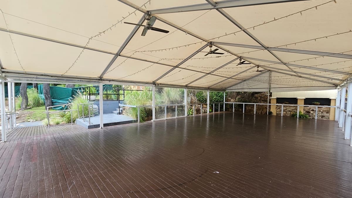 On the left, a step-free ramp with handrails leads to the venue tent. The tent features metal railings and timber floors. Light fixtures and ceiling fans are installed on the ceiling. Adjacent is greenery with wild grass, tall trees, and green mesh.