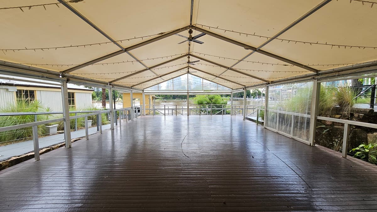 The venue comprises an open space with a pitched tent roof, metal railings, and timber floors. Light fixtures and ceiling fans are installed. To the left is another venue with small windows and a grey corrugated facade. Wild grass, trees, and plants line both sides. Further ahead is a river bordered by metal railings.