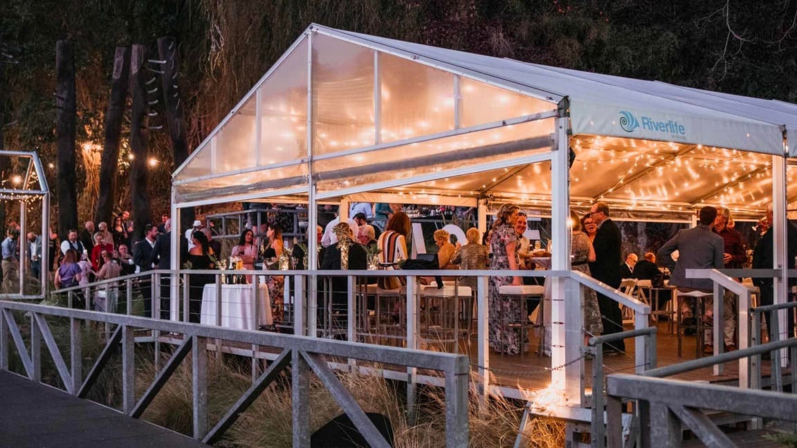 The image shows a lively event venue with guests seated, standing, and moving about. Tables with white linens, wine, and flowers are present. Metal fencing encloses the space, and light fixtures adorn the ceiling. To the left is a road with railings, and behind the tent is a cliff covered with grass and trees. Black posts with light fixtures decorate the area. Trees and wild grass complete the scene.
