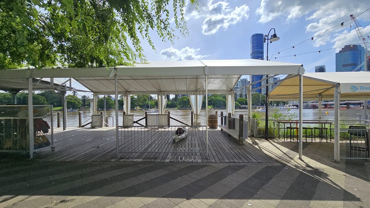 In the centre stands a venue tent with a river view, featuring timber flooring and metal gates for security. A kayak and large barrel display are in the middle, accompanied by a movable storage rack with life vests on the left. Another venue tent is situated to the right, with a tall tree on the left and skyscrapers visible across the river.