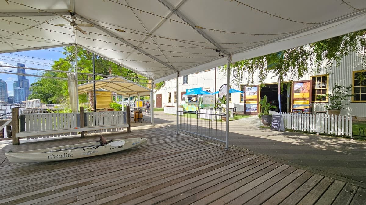 In the centre is a venue tent with timber floors, fenced off by a metal fence and small gates. A kayak is displayed inside. To the right is a facility building with a corrugated facade, small windows, and manual outward-opening doors adorned with colourful posters. In front is a grass area with tables, chairs, large blue umbrella shades, and feather banners. Adjacent is another venue tent with tables and chairs. Trees and skyscrapers are visible in the backdrop.