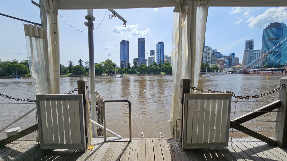 A portion of the venue tent with river views, featuring timber flooring. Metal chain fences and small gates secure the area. Reminder posters are affixed to the gates. White curtains hang at the sides. Across the river, trees and tall skyscrapers, including an iconic bridge, adorn the scenery.