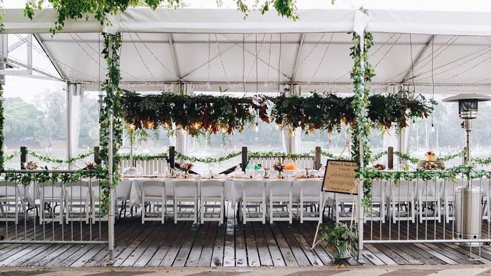 An event venue adorned with vines on metal railings. At the centre sits a long table dressed in white linens, set with cutlery, glassware, and plates. Above, light fixtures illuminate hanging foliage. The floor is timber and there is a lamp post, a rubbish bin, signage, and floral embellishment on the right. The river forms is on the backdrop.