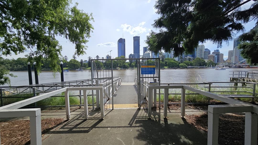 A ramp with a steel gate entrance, handrails, and yellow truncated domes. Warning signage is attached to the gate. Tall black posts are visible nearby. In front, there's a concrete space with metal railings. Trees, wild grass, and metal railings line the area. Across the river, trees, skyscrapers, and an iconic bridge are visible.