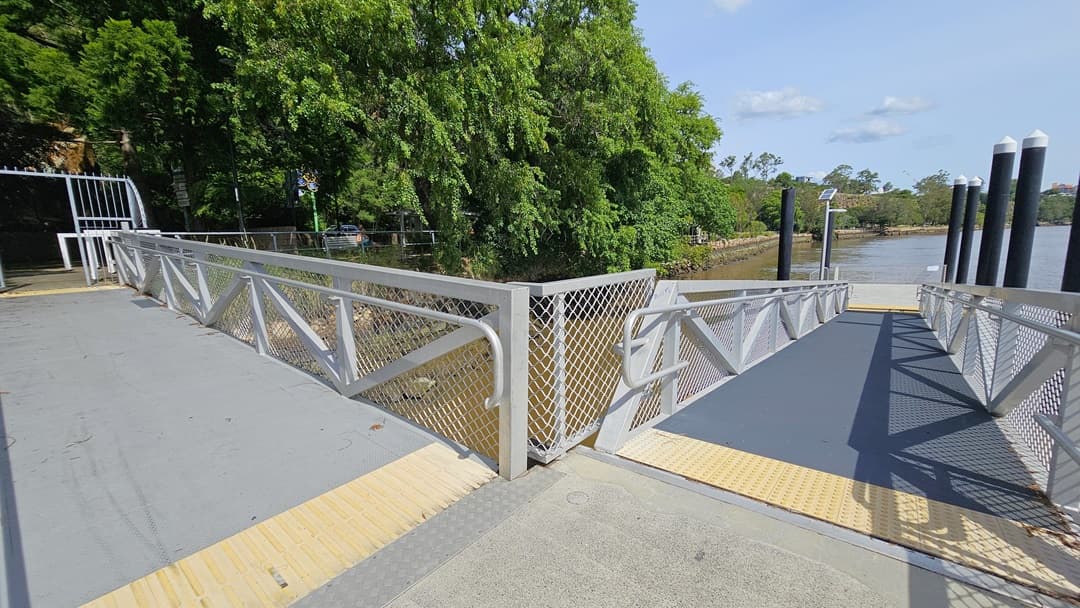 A step-free L-shaped ramp with a steel gate entrance and handrails featuring metal mesh and yellow truncated domes provides access to the riverfront. Nearby, tall black posts support solar-powered light fixtures. The area is bordered by trees, wild grass, and metal railings.