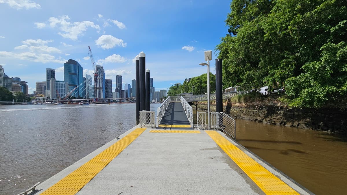 An L-shaped ramp provides step-free access to the riverfront, featuring a steel gate entrance, handrails with metal mesh, and yellow truncated domes. Alongside are tall black posts with solar-powered light fixtures. The ramp extends to an open concrete area bordered by yellow truncated domes. On the right, the area is lined with trees, wild grass, and metal railings, while tall skyscrapers and the iconic bridge grace the left side.