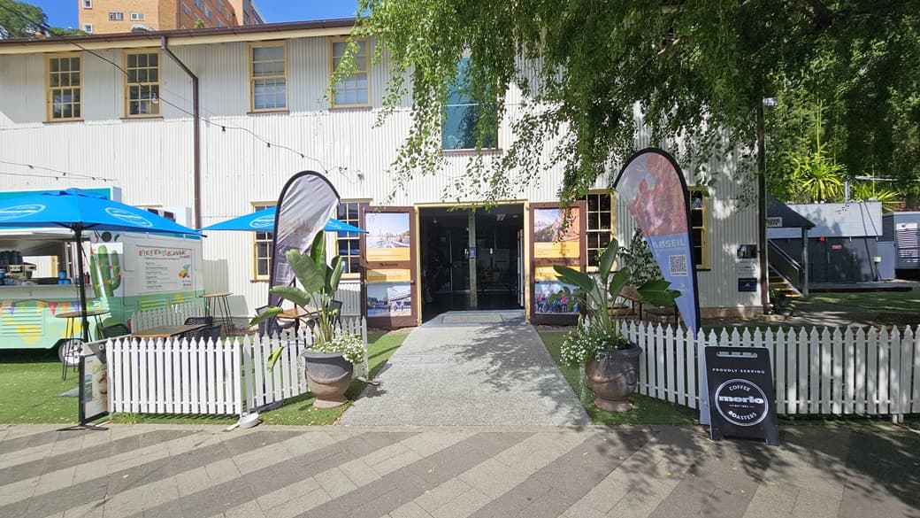 The Riverlife Adventure Centre is a two-storey building with glass windows and a white corrugated facade. It has an outward opening door with activity posters. Outside, tables and chairs sit under a tree. Greenery, including potted plants and grass, adorns the front along with a low white fence, a feather banner, and signage. At the far end, there's a shaded facility with railings.