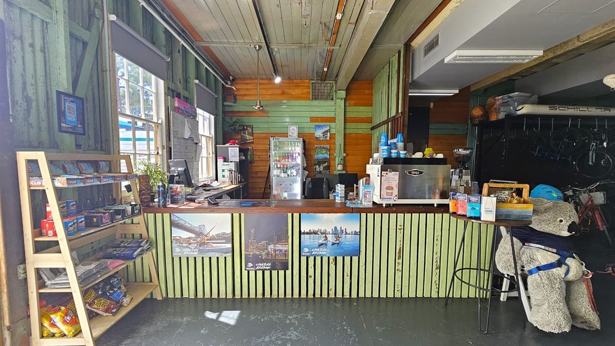 The reception desk has a wooden top and plank front adorned with posters. On it are a beverage dispenser, plastic cups, brochures, a computer, and accessories. A storage rack that holds food items for sale is displayed near the door. A large teddy bear is to the right, with a fridge behind the desk for cold drinks. Bicycles for hire are displayed on the right. Wooden planks decorate the ceiling and walls.