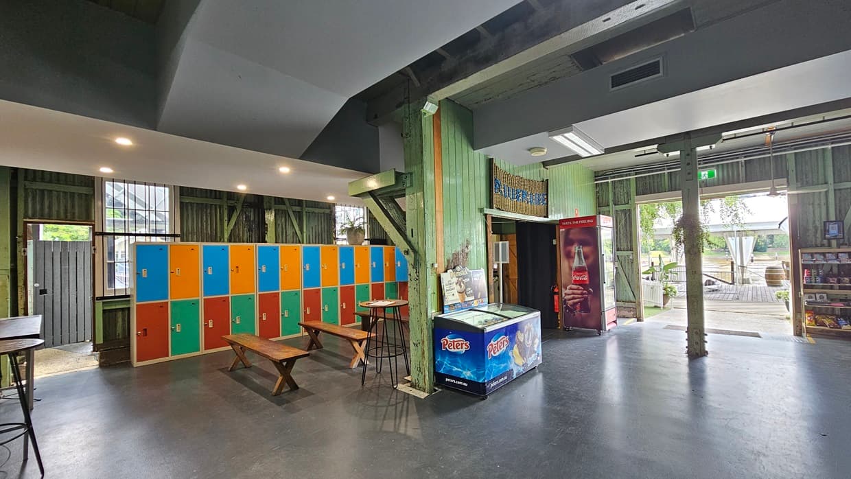 To the left, tables and chairs are arranged in front of colourful lockers. An ice cream freezer is positioned against the middle wall, with a fridge nearby for cold drinks. A banner with the facility name is placed above the door. A storage rack holds food items for sale near the door. The floor is grey, while the wall features green wooden planks. The ceiling is grey with LED lights.