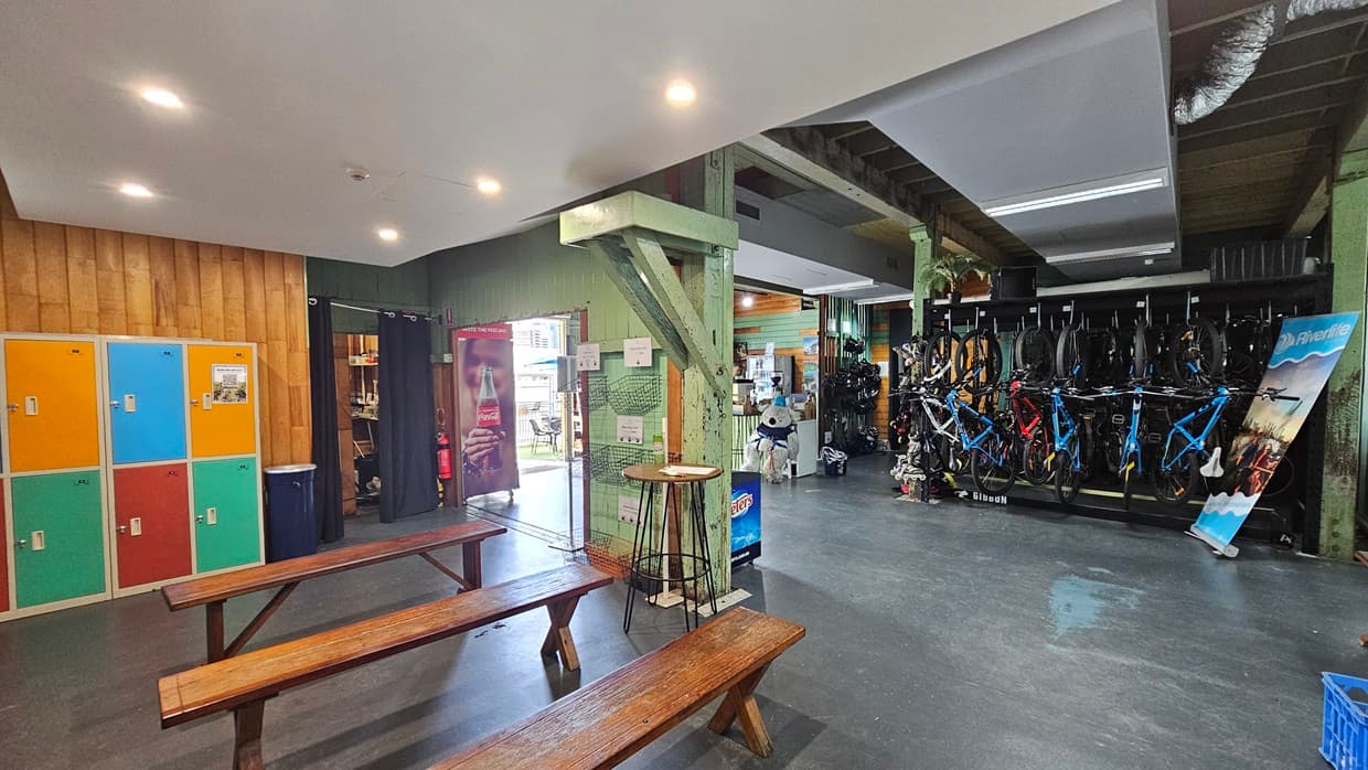 To the left, colourful lockers line the timber wall. Wooden bench chairs and a stool are arranged in the middle, with a nearby rubbish bin. To the right, bikes for hire are displayed. A large retractable poster leans against the green wall. The ceiling has wooden planks and a white surface with LED lights.