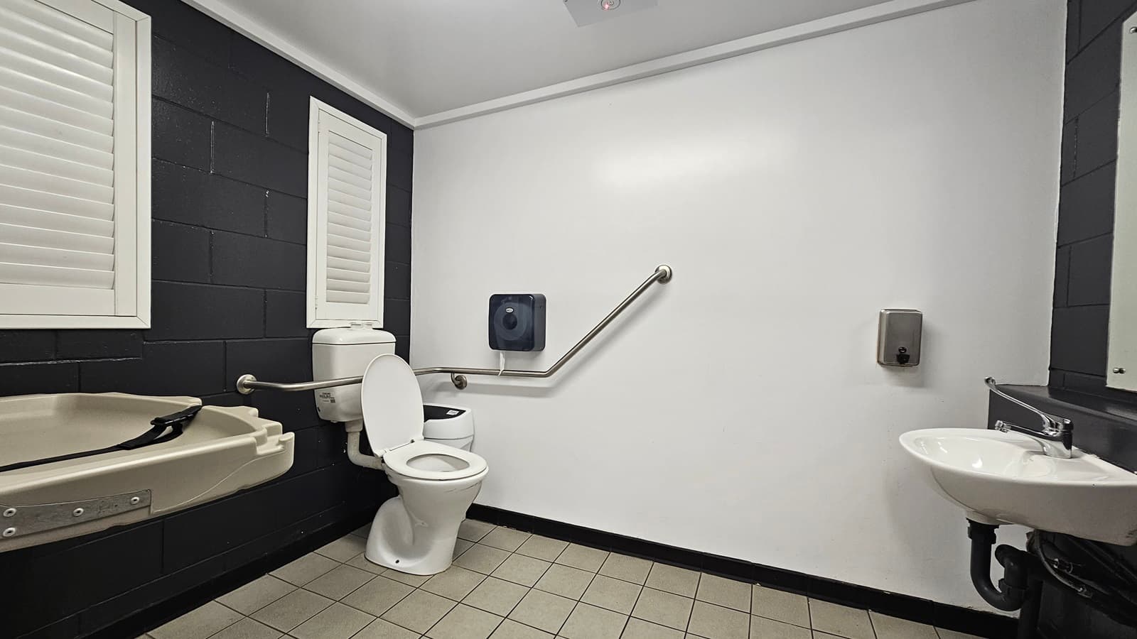 In the corner is a toilet bowl with hand grabs, accompanied by a tissue dispenser and a rubbish bin. Nearby, a change table is available. To the right, a hand basin, liquid soap dispenser, and mirror are mounted on the wall. The room has white walls and ceiling, grey tiled floor, and two small windows with blinds on the black wall to the left.