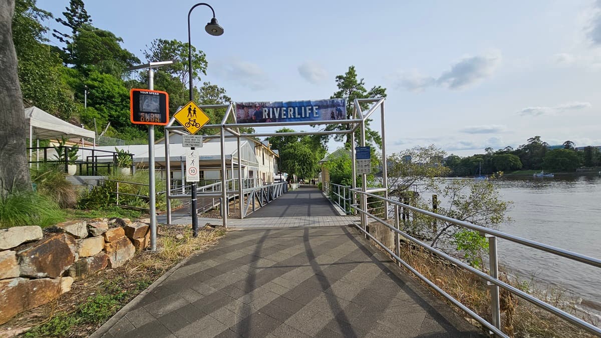 In the centre stands a metal arc displaying the facility name. A lamppost with signages is on the left along with a solar-powered digital signage. Rock formations, wild grass, and trees border the left side, while the Brisbane River is to the right. Metal railings line the road on the right. A ramp with handrails leads to the building. The road has a mix of white and grey bricks.