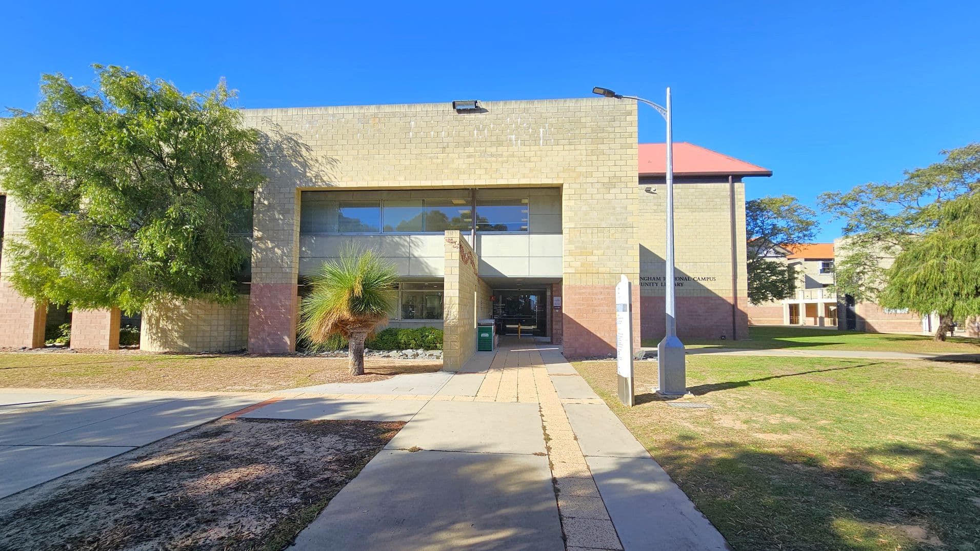 A building with a beige brick facade and large windows in the centre. A pathway leads to the entrance, flanked by a small tree on the left and a lamp post on the right. The building has a flat roof, and there are trees on both sides, with grass and patches of dirt in the foreground. Another building with a red roof is partially visible in the background to the right.